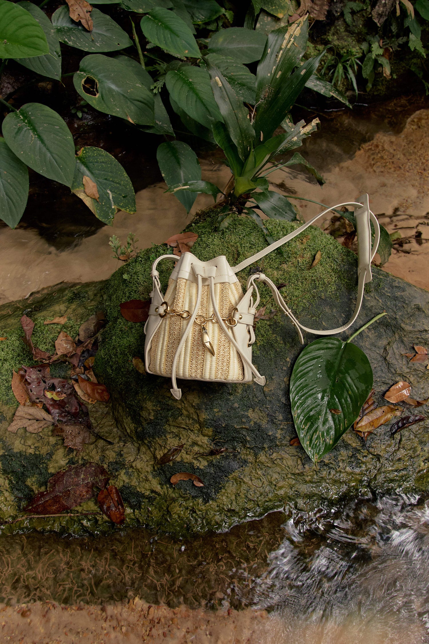 Bolsa bucket bege com detalhes dourados sobre pedra musgosa em ambiente natural, ideal para acessórios sofisticados e funcionais.