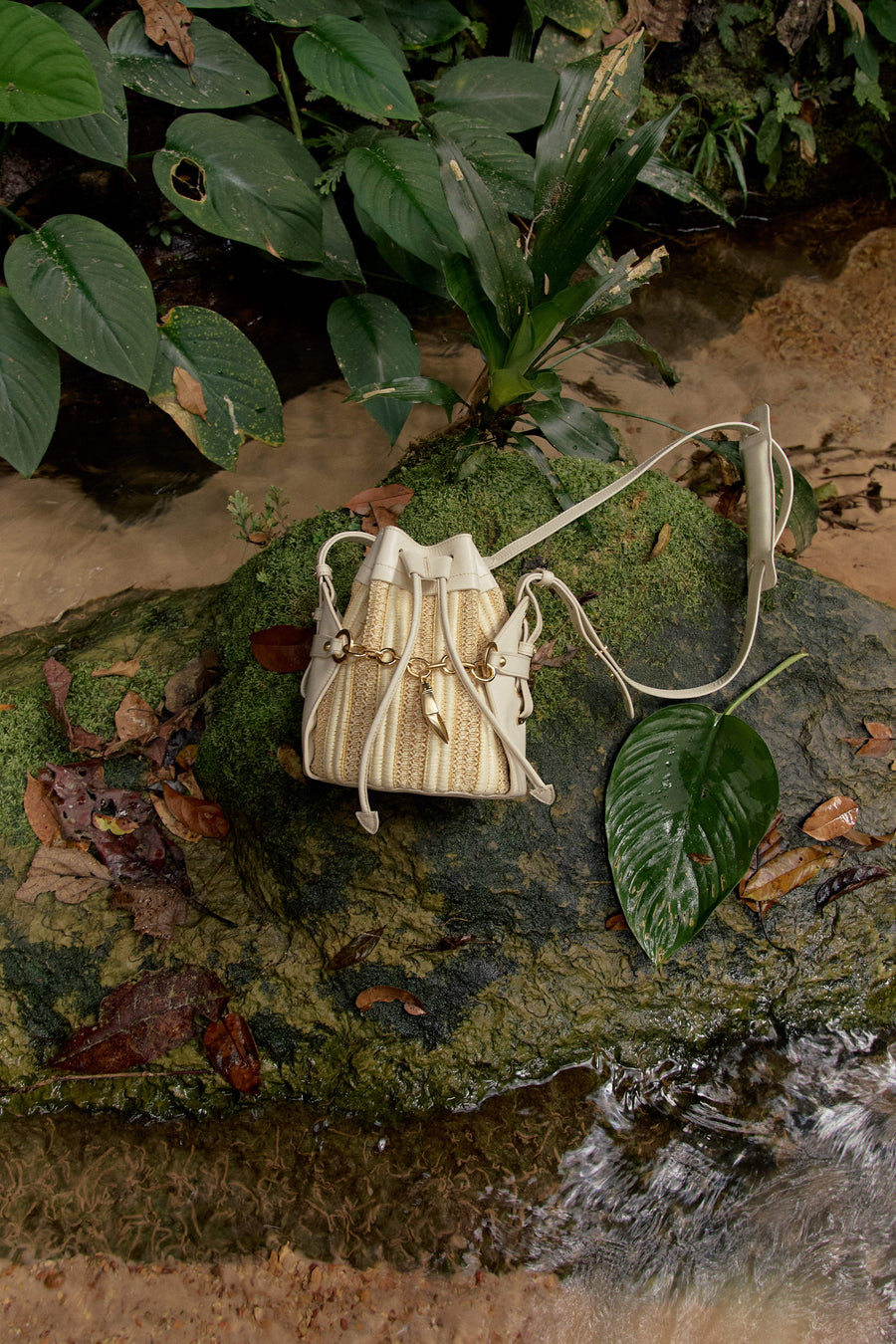 Bolsa bucket bege com detalhes dourados sobre pedra musgosa em ambiente natural, ideal para acessórios sofisticados e funcionais.