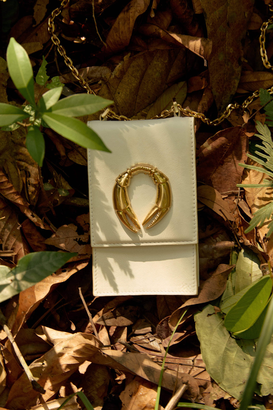 Bolsa branca com detalhe dourado em forma de garras, sobre folhas secas e plantas verdes