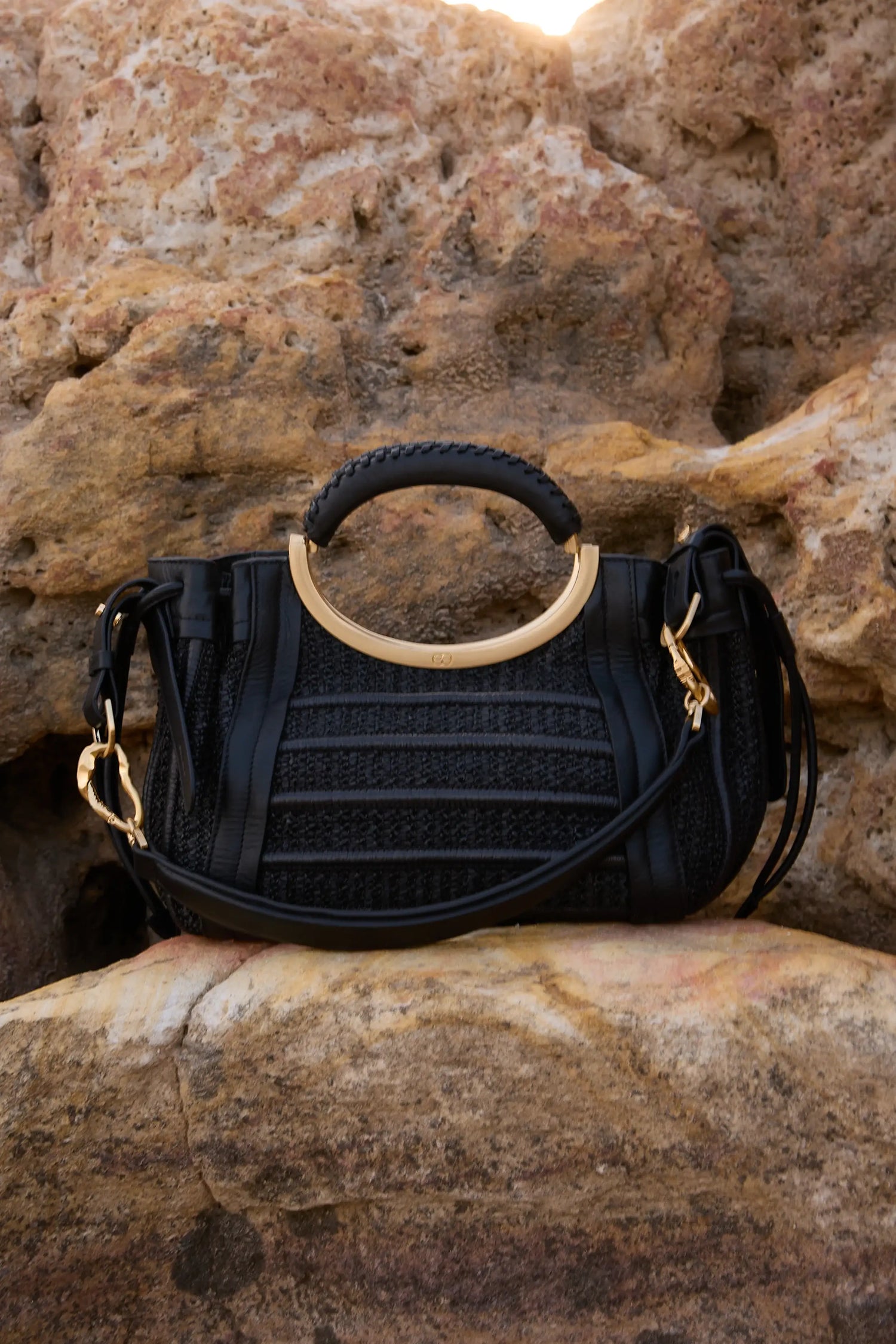 Bolsa preta com alça circular dourada e detalhes em couro, sobre rochas ao pôr do sol