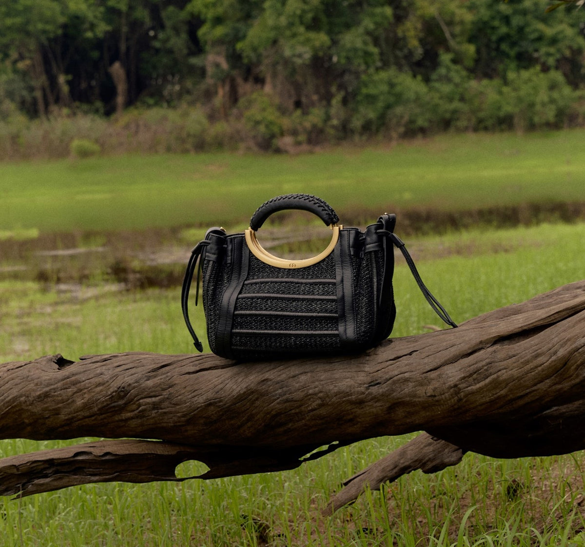 Bolsa preta com alça circular dourada sobre tronco em paisagem verde, design brasileiro sofisticado