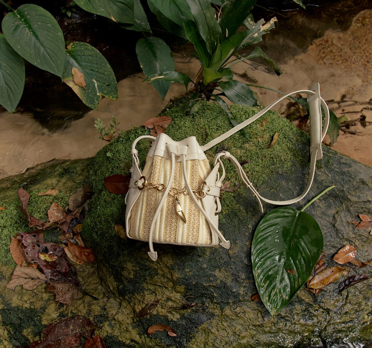 Bolsa bucket bag em tons neutros com detalhes em dourado, sobre pedra musgosa em ambiente natural com folhagens