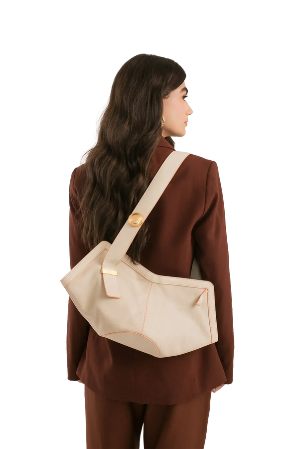 Bolsa off-white com costura rosa, alça larga e detalhe dourado, usada por mulher de blazer marrom.