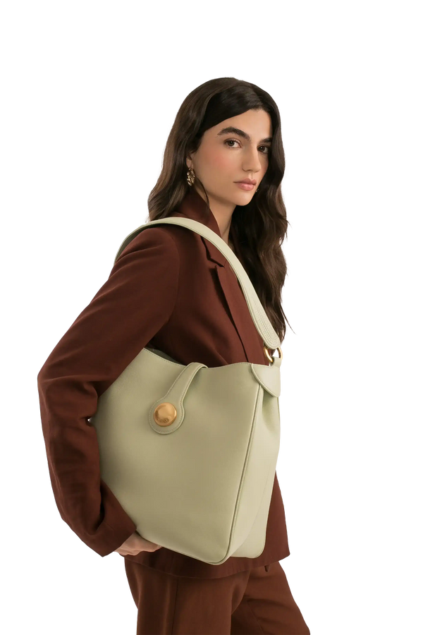 Bolsa verde claro com alça larga e detalhe dourado, usada por mulher em blazer marrom.