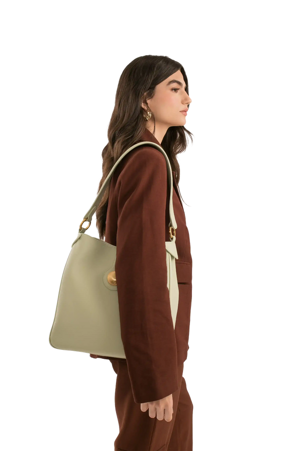 Bolsa verde claro com alça longa e detalhes dourados, usada com blazer marrom. Design brasileiro e sofisticado.